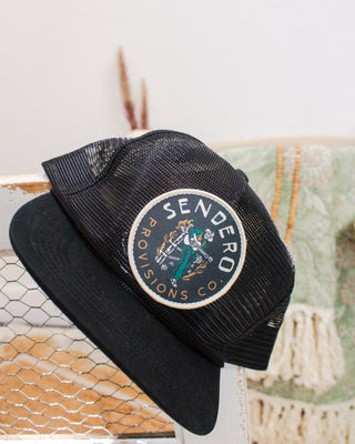 Black mesh trucker hat from Sendero Provisions Co. with a round patch featuring a cowboy illustration, displayed on a rustic backdrop with neutral decor.