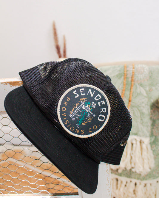 Black mesh trucker hat from Sendero Provisions Co. with a round patch featuring a cowboy illustration, displayed on a rustic backdrop with neutral decor.