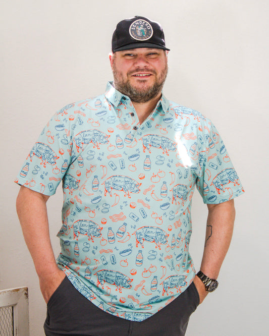 Man wearing a light blue short-sleeve button-up shirt with a colorful BBQ-themed print, including pigs, bacon, hot sauce, and tomatoes, styled with a black Sendero hat.