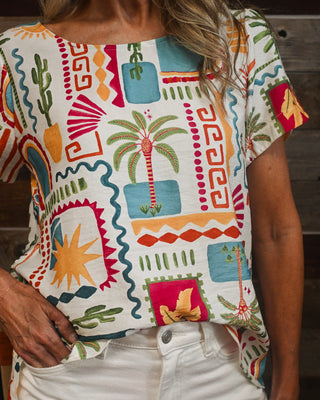 Women's colorful tropical graphic print blouse featuring palm trees, cactus, sun and geometric patterns, styled with white denim for a vibrant resort-inspired boutique outfit.