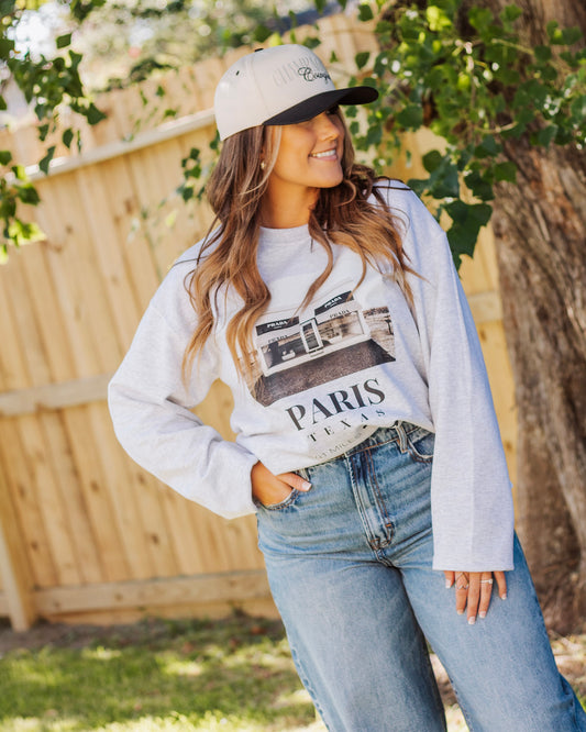Smiling woman in a gray Paris Texas graphic sweatshirt with Prada storefront print, paired with relaxed blue jeans and a beige trucker hat, casual street style.