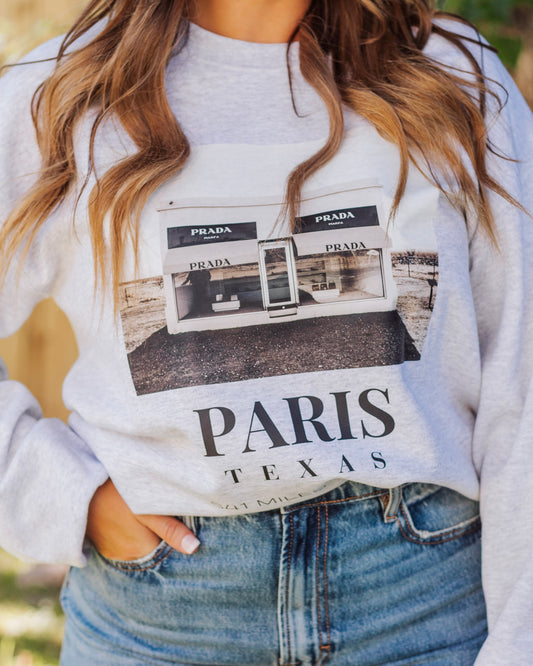 Woman wearing a light gray Paris Texas graphic sweatshirt with Prada storefront print, paired with high-waisted blue jeans for a casual chic outfit.