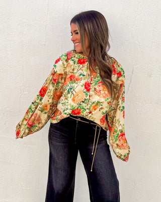 Woman wearing a floral satin blouse with puff sleeves and tie neckline in red, peach, and green tones, styled with dark high-waisted jeans for a chic fall look.