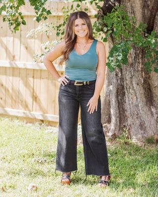 Woman wearing a teal sleeveless tank top tucked into high-waisted black wide-leg jeans with a gold buckle belt, styled with gold layered necklaces for a polished look.