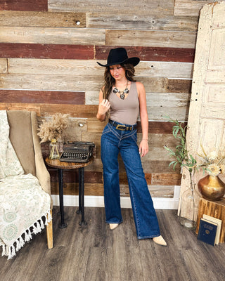 Women's western chic outfit featuring a taupe sleeveless tank top, dark wash high-waisted wide-leg jeans, black cowboy hat, statement necklace, western belt and cream pointed-toe boots.