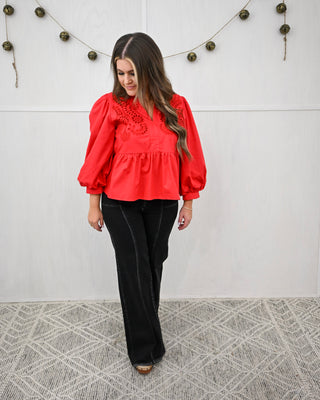 Woman wearing a red blouse with lace details and black pants against a white wall.