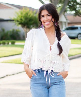 Woman wearing a white embroidered eyelet tie-front blouse with scalloped hem and puff sleeves, paired with high-rise light-wash denim. A feminine and breezy look perfect for spring and summer.