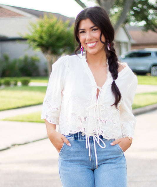 Woman wearing a white embroidered eyelet tie-front blouse with scalloped hem and puff sleeves, paired with high-rise light-wash denim. A feminine and breezy look perfect for spring and summer.