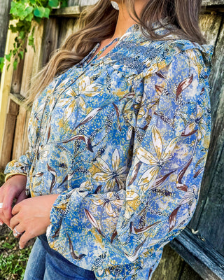 Blue floral blouse with smocked shoulder detail and ruffle trim, featuring a mix of yellow, cream, and brown blooms for a feminine, boho-chic boutique look.