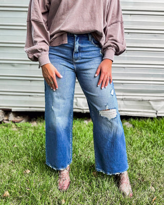 Woman wearing distressed wide-leg jeans in medium wash with raw hem, paired with a mauve long sleeve top and snakeskin booties for a chic boutique look.