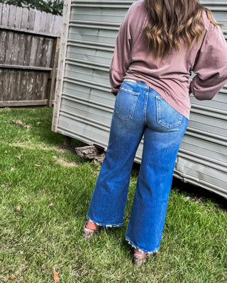 Woman wearing high-rise medium wash wide-leg jeans with frayed hem, styled with a mauve long sleeve top for a relaxed boutique denim look.