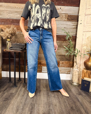 Women's medium wash wide leg jeans with subtle fading and raw hems, styled with a charcoal graphic tee and nude pointed toe flats at MaKenzie Layne Boutique.