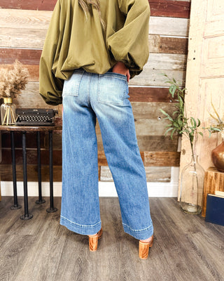 Women’s high-rise wide leg jeans in a medium wash with back patch pockets and relaxed full-length fit, styled with an olive green blouse and heels.