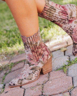 Side view of women’s floral tapestry ankle boots with stacked wooden block heel and pointed toe, styled outdoors on brick walkway for a vintage western look.