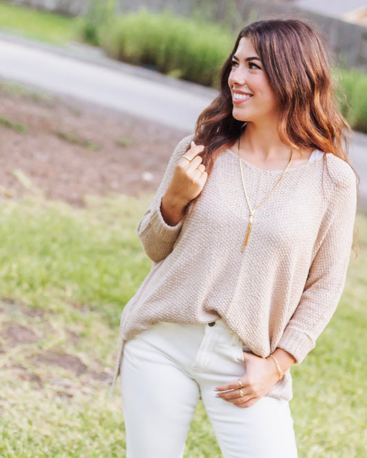 Woman in a beige knit sweater paired with white high-rise jeans, accessorized with a long gold necklace and rings, standing outdoors with a casual chic look.