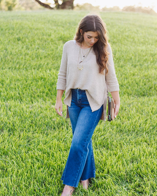 Woman wearing a beige knit sweater with side slits, paired with high-waisted flared blue jeans and layered gold necklaces, standing on a grassy field.