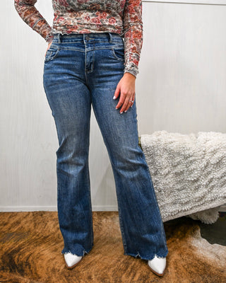 Person wearing blue jeans and a patterned top in a room with a white wall and textured rug.
