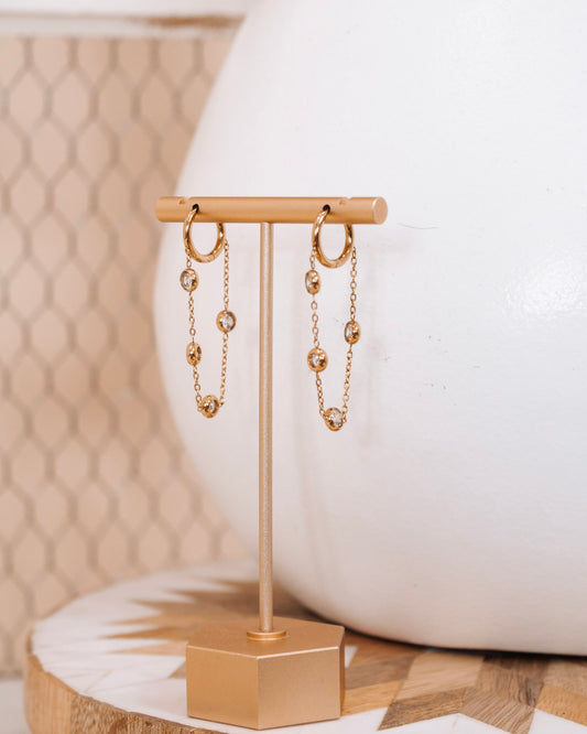 Pair of gold hoop earrings with hanging crystal-accented chains displayed on a modern gold stand against a neutral background with geometric and ceramic decor.