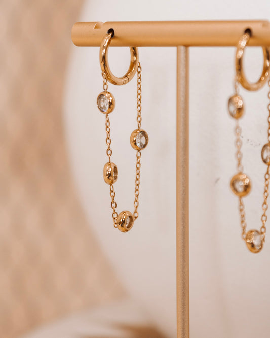 Gold hoop earrings with dangling round crystals on a delicate chain, displayed on a gold earring stand against a soft neutral background.