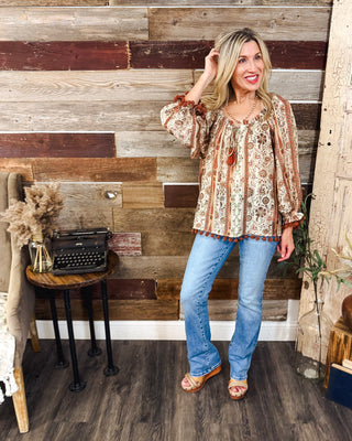 Women’s boho floral print blouse in warm fall tones with long sleeves and rust pom-pom trim, styled with light wash flare jeans for an easy autumn outfit.