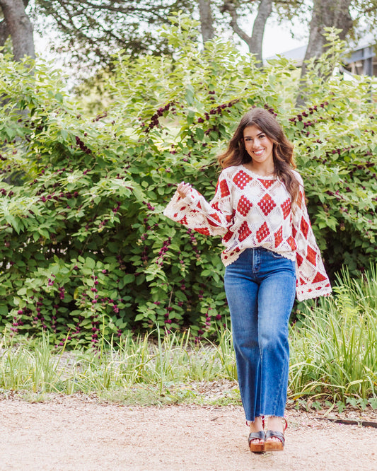Woman wearing a boho crochet sweater with red and cream diamond patterns, paired with high-rise blue jeans and platform sandals, standing outdoors by greenery.