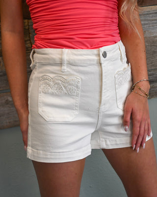 Women’s white high-rise denim shorts with lace trim front pockets and cuffed hem, styled with a bright pink top for a feminine boutique look