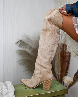 Close-up of a tall beige suede western boot with stitched embroidery, stacked block heel, and pointed toe, styled with a plaid shirt and tiered lace mini skirt.