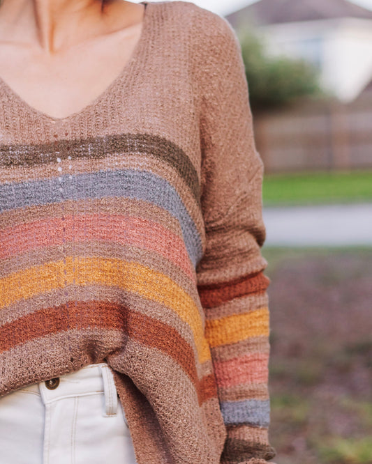 Close-up of a cozy knit sweater with multicolor horizontal stripes in earthy tones of brown, mustard, orange, rust, and blue, styled with white denim jeans.