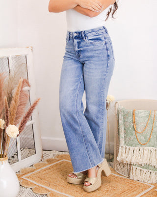 Front view of a woman wearing high-rise light wash jeans with a subtle flare and raw hem, paired with a white tank top and platform sandals.
