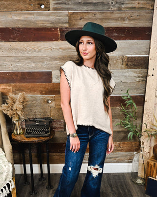 Cream sleeveless sweater vest with black whipstitch trim and relaxed fit, styled with distressed dark wash flare jeans for a chic western-inspired outfit.