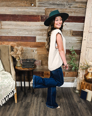 Cream sleeveless sweater vest with contrast whipstitch trim, styled with dark wash flare jeans and a wide brim hat for an effortless western fall look.