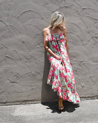 Women's tropical floral maxi dress with pink hibiscus-inspired print, ruffle neckline, adjustable straps, and flowing tiered hem, styled with wedge sandals for a resort-ready summer boutique look.