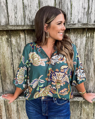 Woman wearing a dark teal floral blouse with puff sleeves and a relaxed fit, featuring earthy tones of tan, olive, and cream, paired with dark denim jeans.