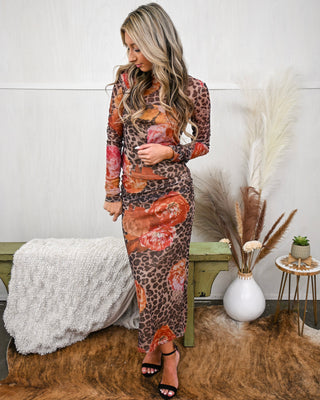 Woman wearing a long-sleeve leopard and floral print mesh bodycon maxi dress with ruched sides, styled with black heels and loose curls in a boutique setting.