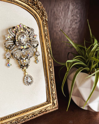 Ornate vintage-inspired crystal brooch with iridescent rhinestones, antique gold-tone detailing and dangling teardrop accents for statement styling.