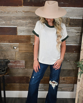 Women’s ivory micro stripe short sleeve top with dark green contrast trim, styled with distressed dark wash jeans and a neutral wide brim hat for a casual fall outfit.