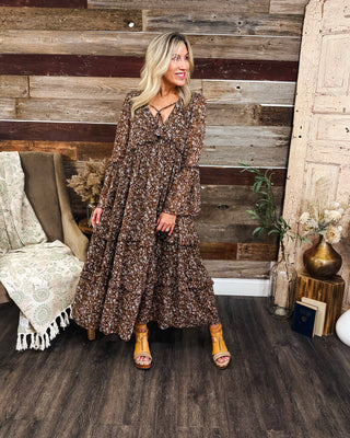 Women's brown ditsy floral maxi dress with a flowy tiered skirt, long bell sleeves, and tie-front V-neckline, styled for a boho western fall look at MaKenzie Layne Boutique.