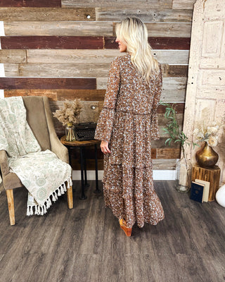 Back view of women's brown ditsy floral print maxi dress with long bell sleeves, a tiered flowy skirt, and boho western style at MaKenzie Layne Boutique.