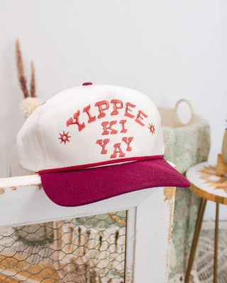 Cream and maroon trucker hat with “Yippee Ki Yay” embroidered in red lettering, displayed on a rustic wooden frame in a Western-inspired setting.