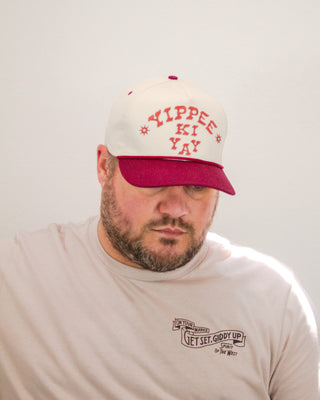 Man wearing a cream and maroon “Yippee Ki Yay” trucker hat paired with a light beige Sendero Provisions Co. t-shirt featuring “Get Set, Giddy Up” chest graphic.